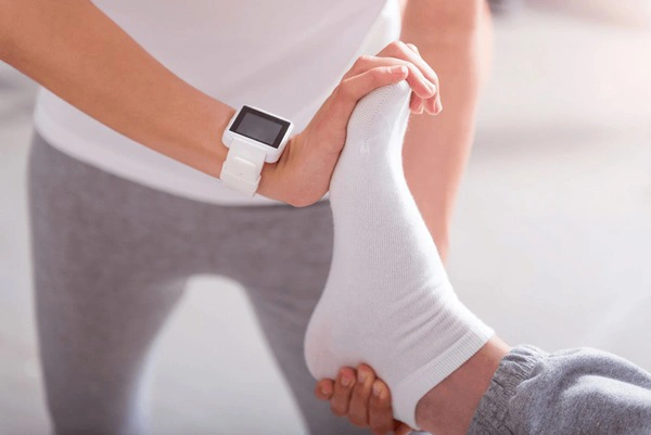 A person in athletic clothing holds the foot of another person wearing a white sock, indicating a physical therapy session.